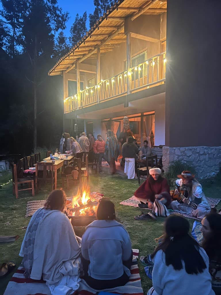 People gathered around a bonfire on the lawn at night with guitar music and the house balcony lit with string lights