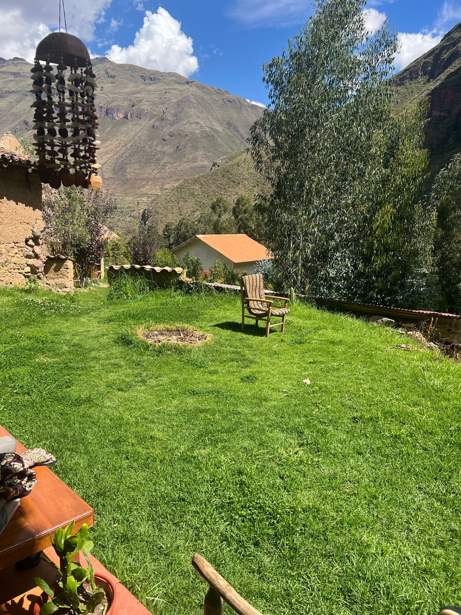 A lawn with a mountain backdrop, a chair near the center, and tall trees beside the house